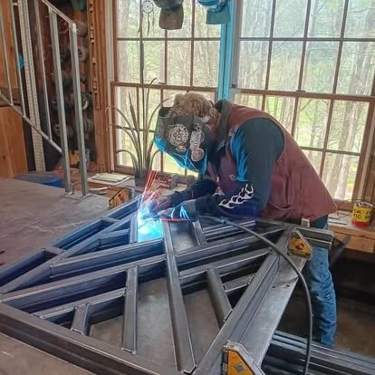 A person wearing protective gear welding a metal frame inside a workshop with large windows and wooden walls.