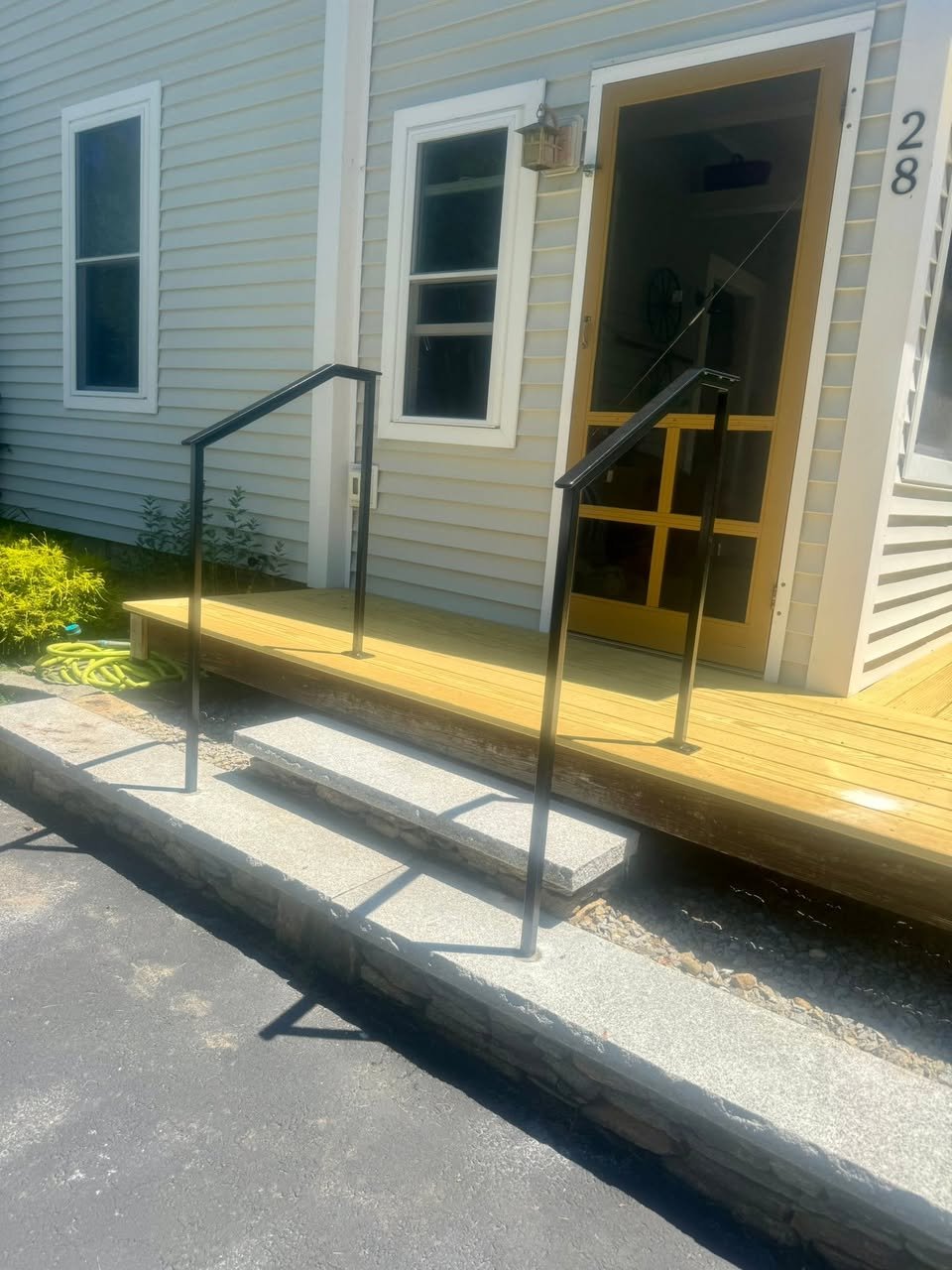 Front porch with a small wooden ramp and black metal handrails leading to a screened door of a house with light-colored siding and two windows.