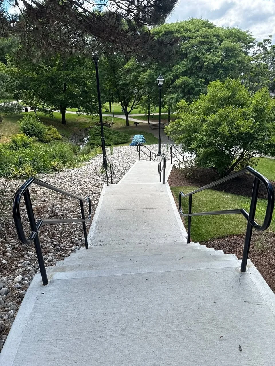 Concrete stairs with black metal handrails leading down to a park area with trees, grass, benches, and lampposts.
