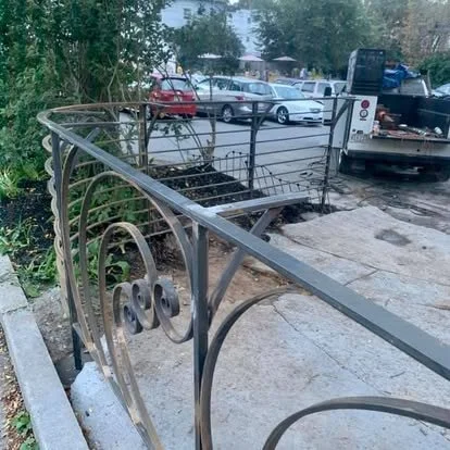 A decorative outdoor metal railing on a sidewalk next to a parking lot with cars and trees in the background.
