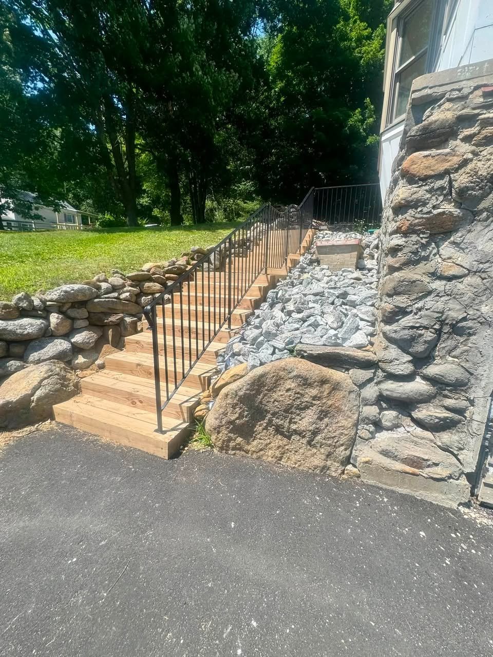 Wooden staircase with black metal railing leading up to a house, with rocky landscape and stone wall on the side, surrounded by trees.