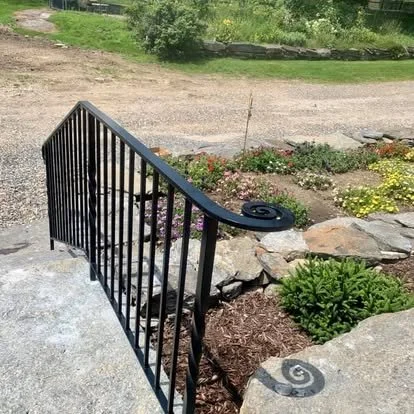 Close-up of a black metal railing on a small outdoor staircase next to a garden with flowers and rocks.