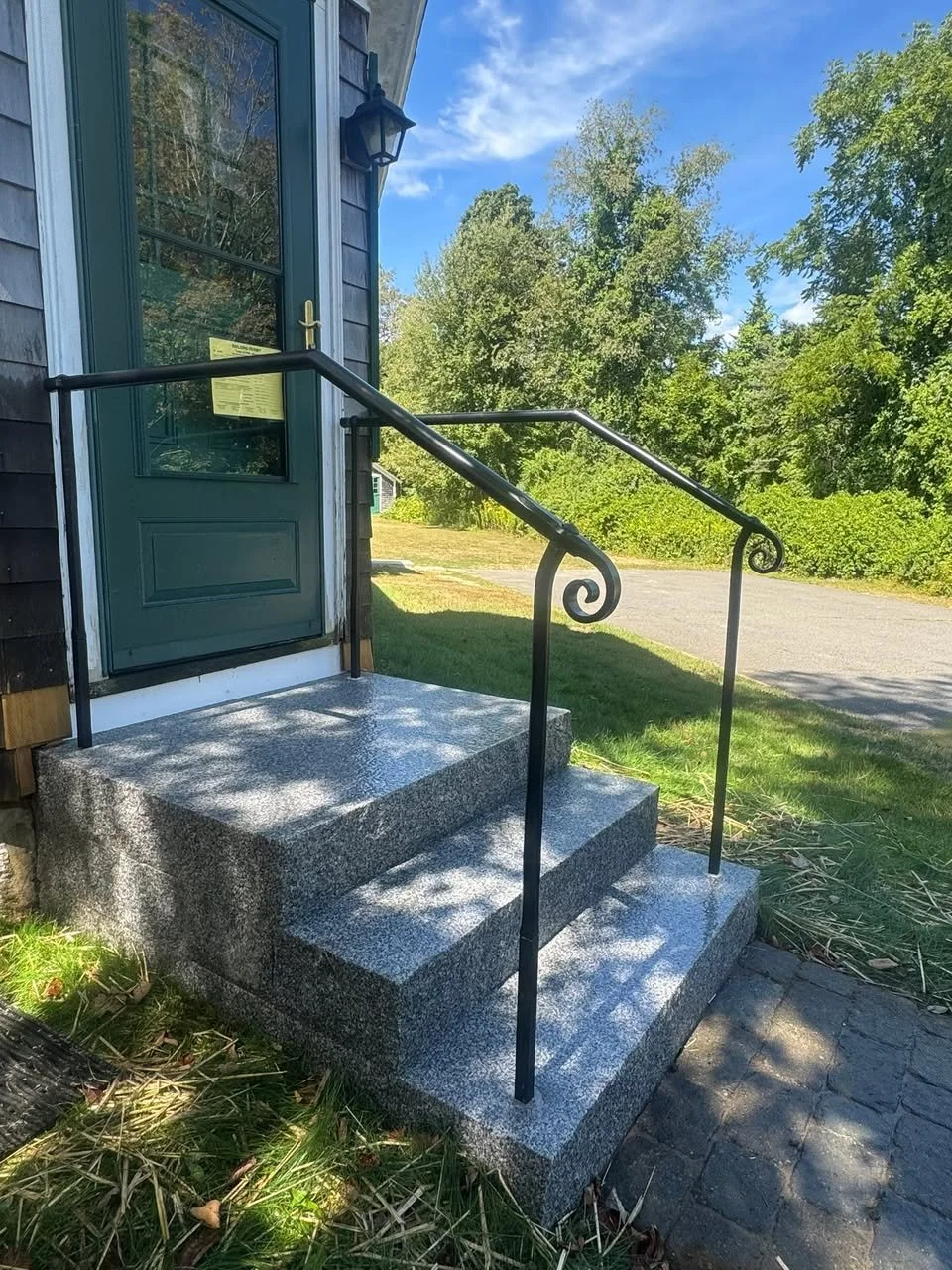 Stone steps leading up to a green front door with black iron railings on either side, outside of a house with dark wooden siding and a black lantern beside the door. The area around the steps has grass and some dried leaves, with trees and a blue sky