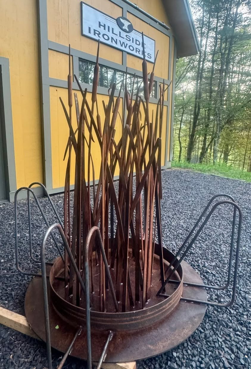Outdoor sculpture of rusted metal stakes arranged upright in a circular base, leaning against a yellow building labeled 'Hillside Ironworks' with a wooded area in the background.