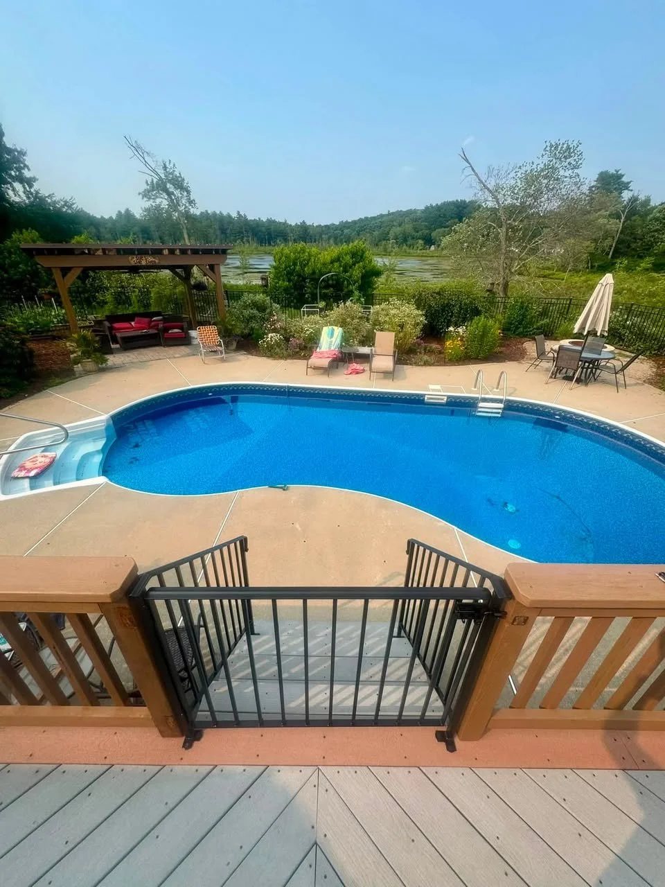 Custom made railing and gate for swimming pool in backyard with lounge chairs, patio table with umbrella, garden, and view of lake and trees in the distance.