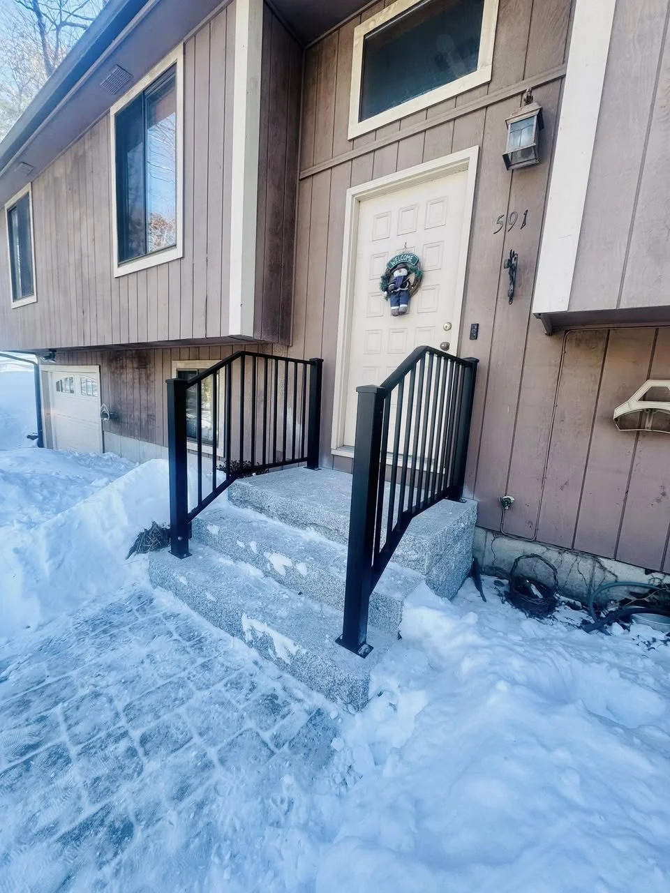 Front entrance of a house with snow-covered steps and a black metal railing. The house has beige wood siding, a white door with a holiday wreath, and several windows. Snow is on the ground and steps.