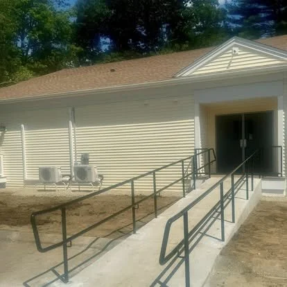 Accessible ramp leading to building entrance with black metal handrails and two air conditioning units outside.