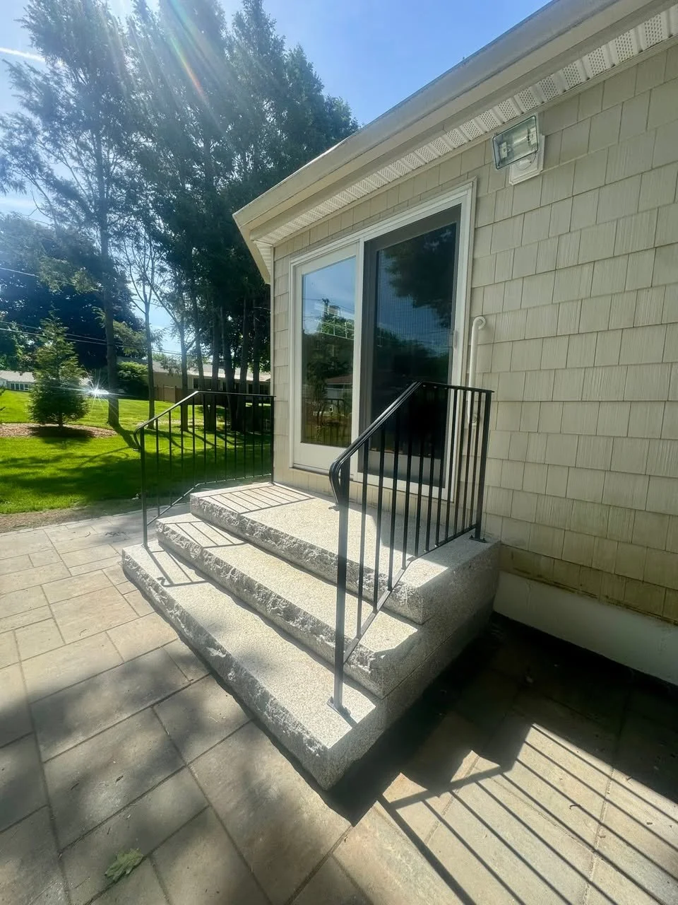 Exterior back door with sliding glass door leading onto a small concrete porch with steps, black metal railing, and a beige siding house under a sunny blue sky.