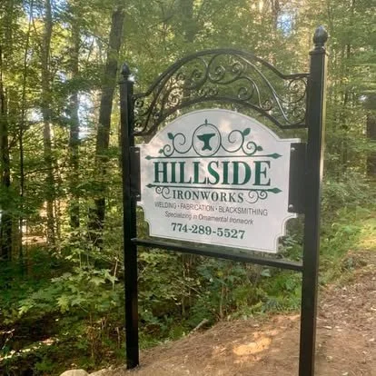 Sign for Hillside Ironworks in a wooded area, advertising welding, fabrication, and blacksmithing services, with a phone number.