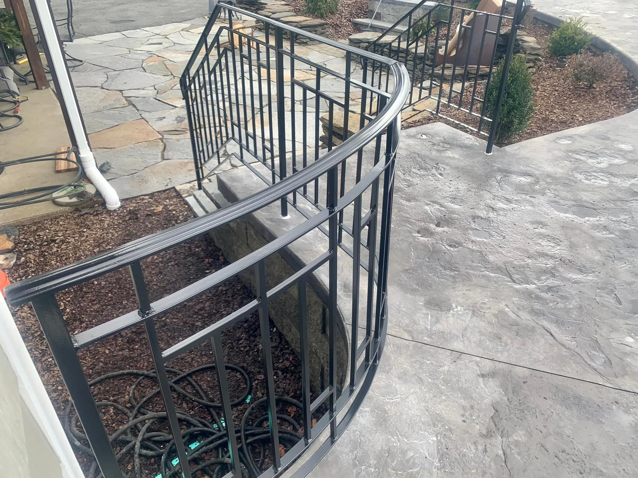 A custom metal staircase with black rails and a curved top step, under construction on a concrete and stone patio, with soil and plants nearby.