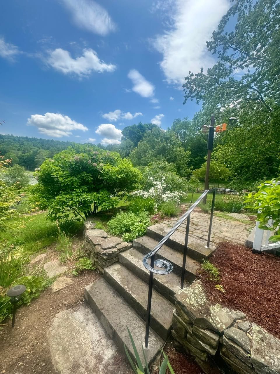 Stone steps with a custom metal handrail in a lush garden under a blue sky with scattered clouds.