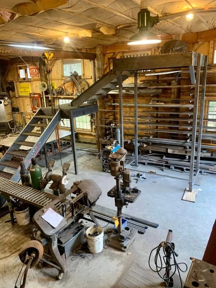 Interior of a workshop with vintage machinery, a staircase leading to a raised platform, and various tools and equipment.