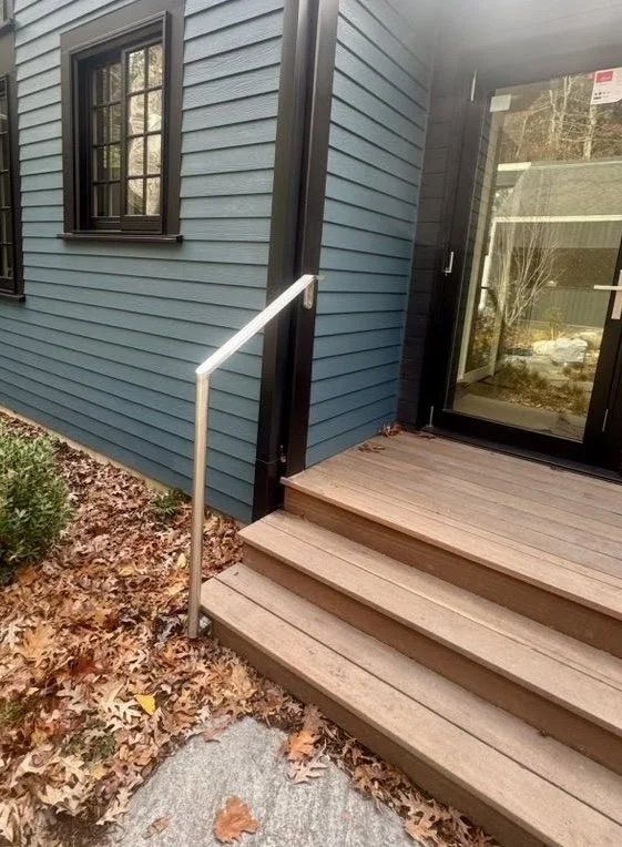 A small wooden deck with stairs leading to a glass door on a blue house, with a metal handrail on the side and fallen leaves on the ground.