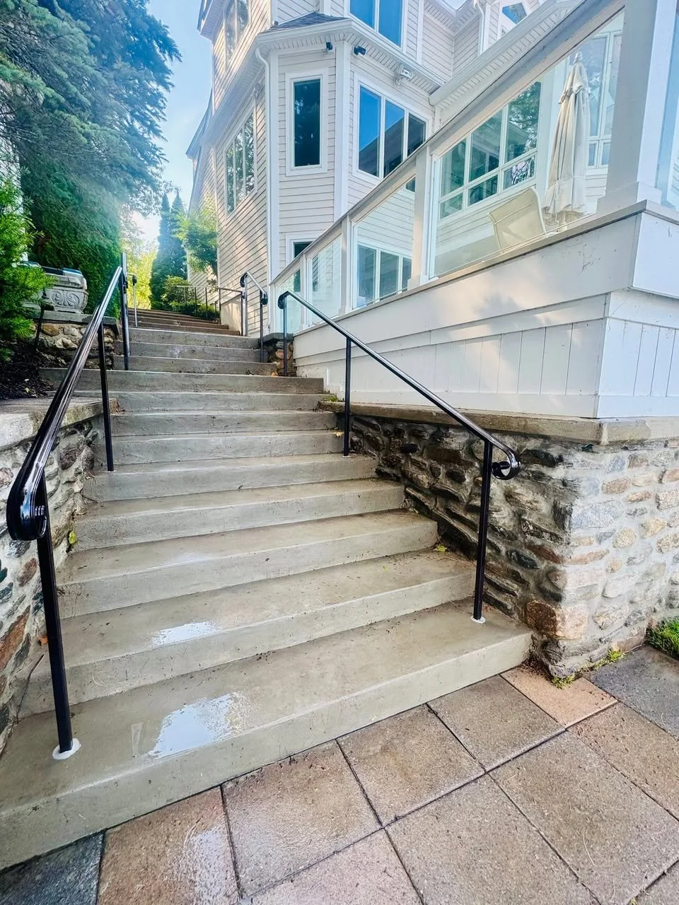 Concrete stairs leading up to a white house with a wrap-around porch and large windows, surrounded by trees and greenery.