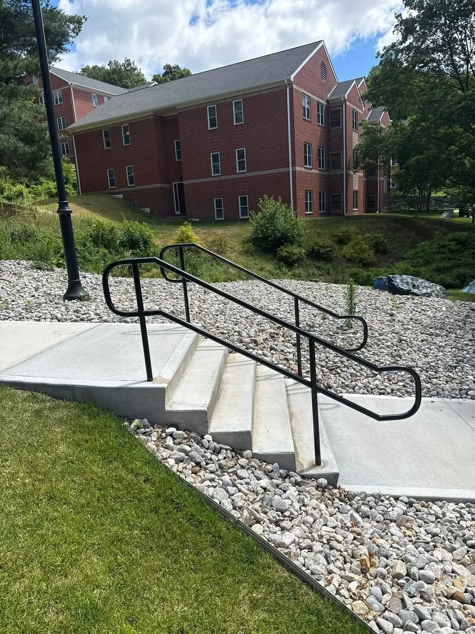 Concrete stairs with handrails on a sidewalk next to grass and gravel, leading up to a landscaped area with bushes and a street lamp. In the background, there is a multi-story brick residential building surrounded by trees.