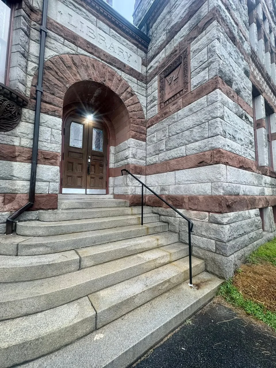 Stone steps lead up to a dark wooden door on the stone exterior of a building. The building has a sign that reads 'LIBRARY' above the archway, with additional inscriptions nearby. A metal handrail runs along the steps on the right side.