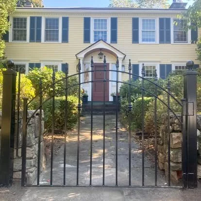 Yellow two-story house with blue shutters and a red front door, accessed by a stone pathway and iron gate, surrounded by greenery.