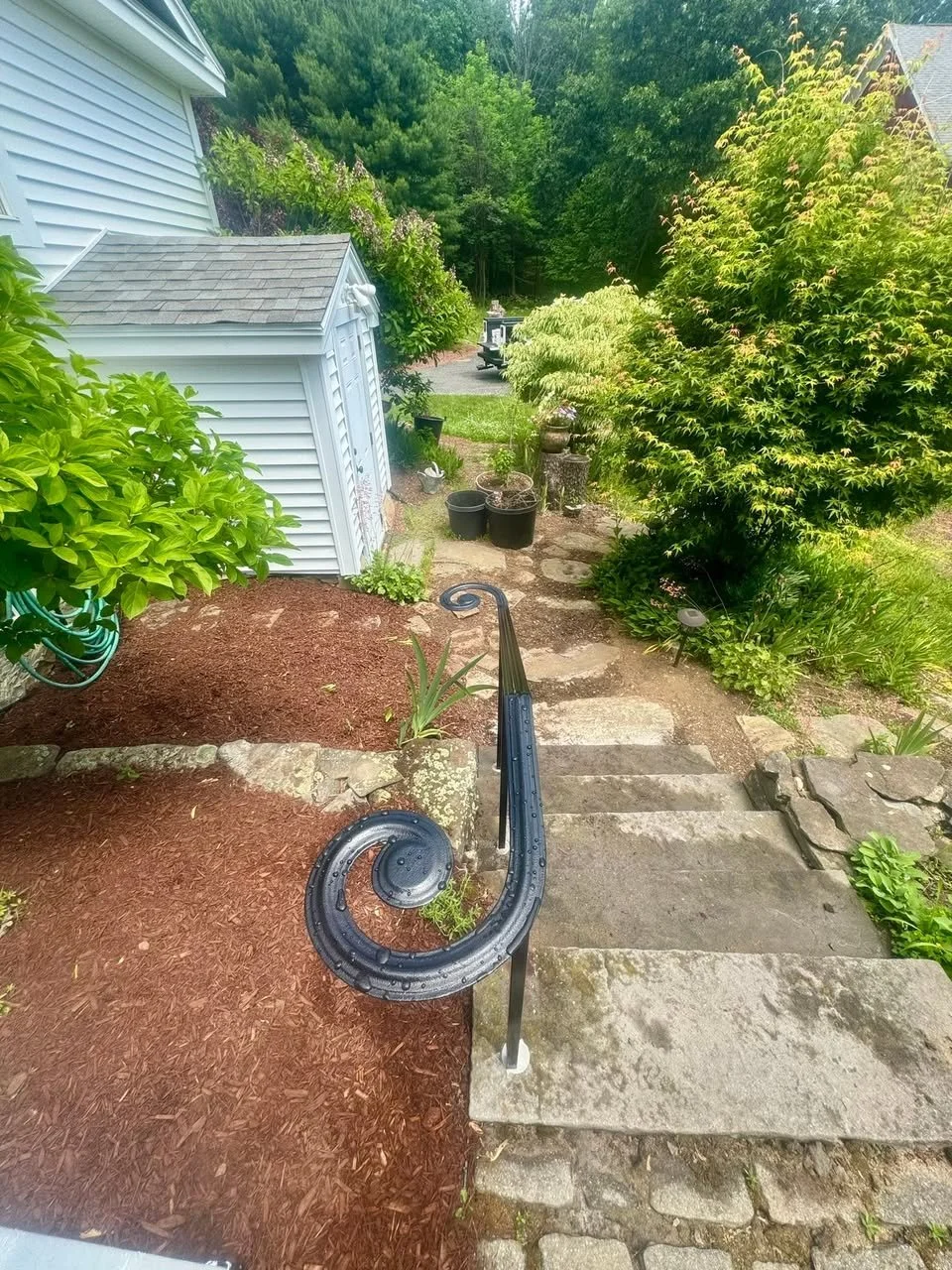 Stone pathway with a black metal handrail leading down steps in a garden with lush green bushes and trees, and a white storage shed on the left, in a backyard with a grassy area and wooded background.