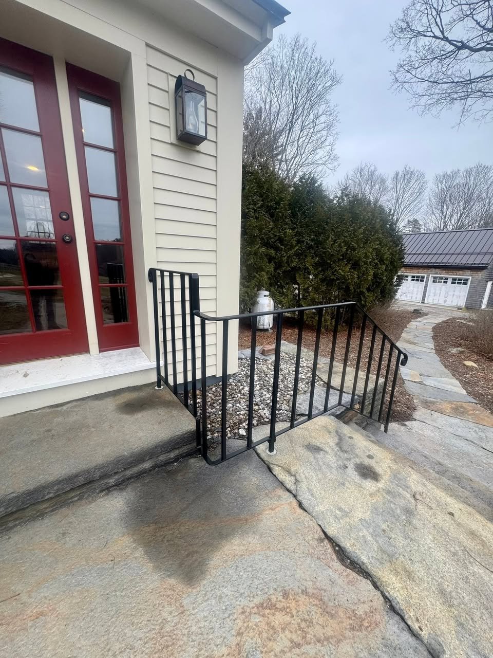 Exterior view of a house with a small ramp and black metal railing leading to a door with glass panels. There is a wall-mounted lantern and a pathway with stones leading to a garage in the background.