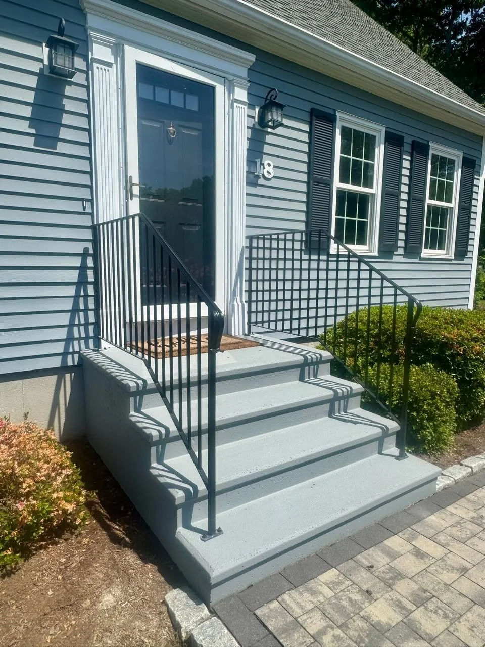 Front porch of a house with gray siding, black front door, white trim, three steps with black metal railings, two black outdoor lanterns, and windows with black shutters.