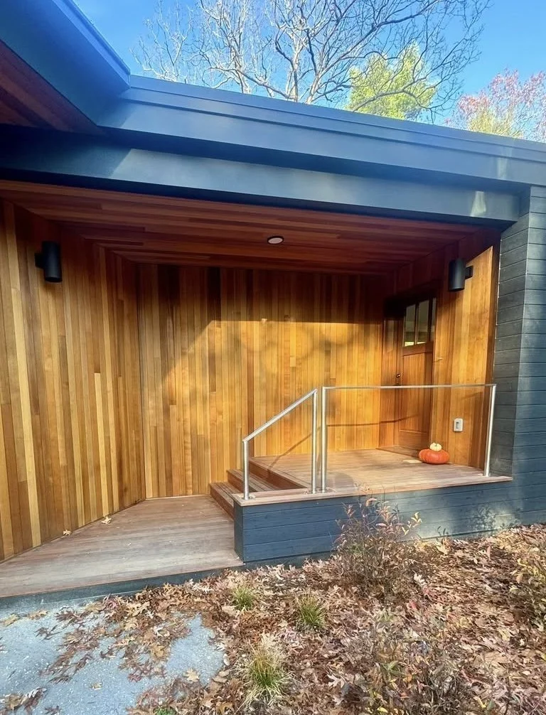 Small porch with wooden panel walls, a wooden floor, a metal railing, and a pumpkin decoration on the floor. The house exterior is painted dark gray with a blue roof. There are fallen leaves and plants in front.