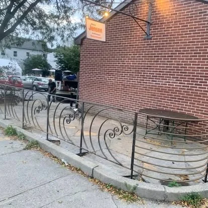 Outdoor patio with a wrought iron railing, a round table with metal chairs, a brick building with an orange sign, and trees in the background.