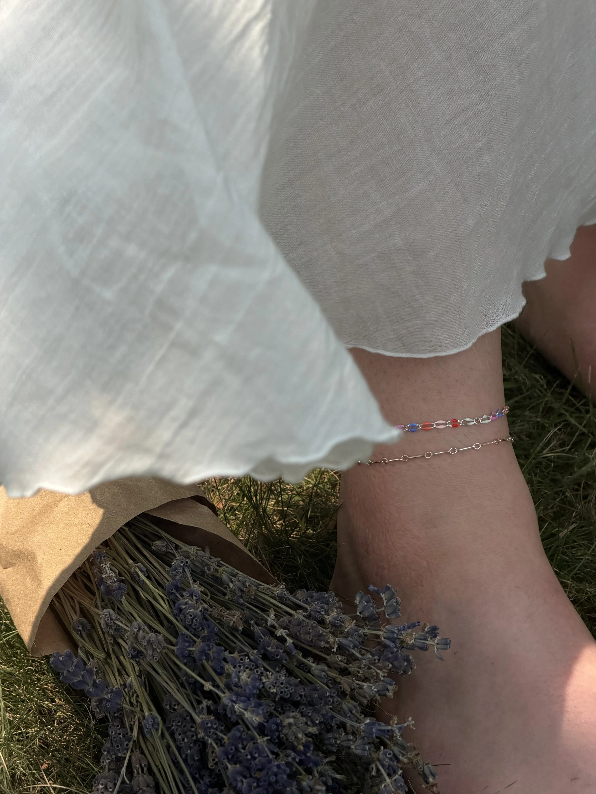 Close-up of a person's leg with a colorful anklet, next to a bundle of lavender flowers in a paper bag, and a light-colored fabric garment partially covering the scene, outdoors on grass.