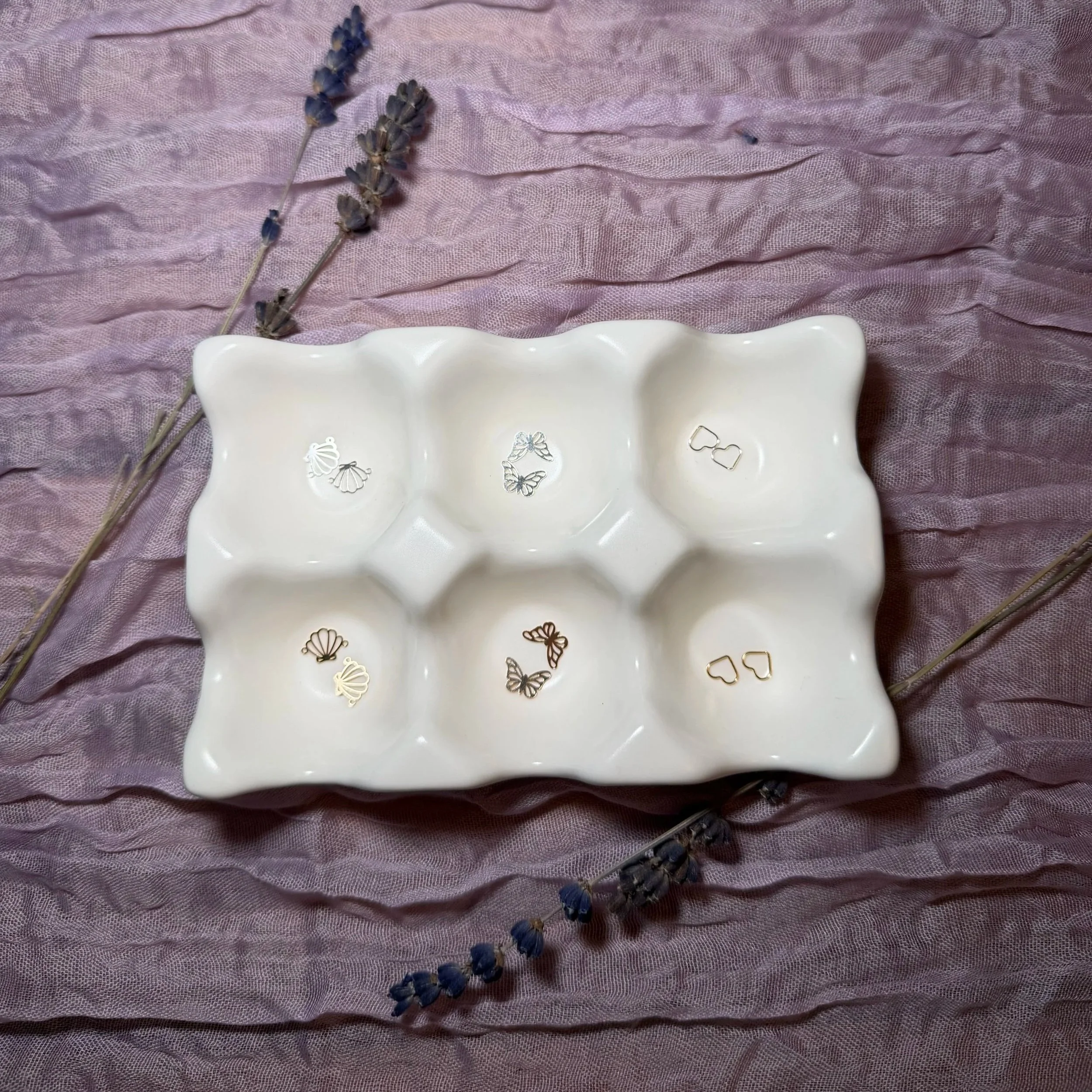 Two sprigs of dried lavender flowers placed on a pink textured fabric background, partially overlapping a white ceramic jewelry dish containing various small jewelry pieces.