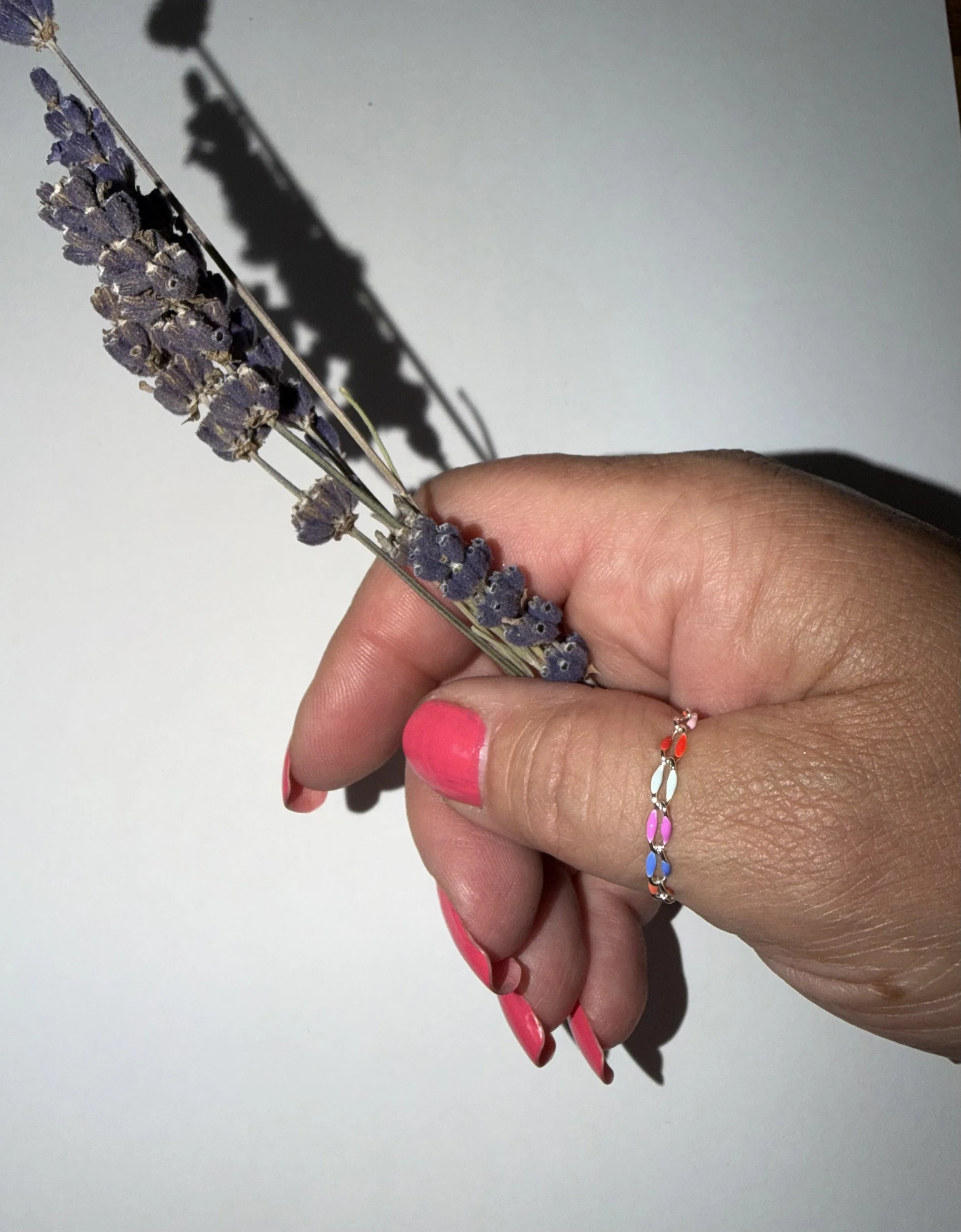 A hand holding a lavender sprig with purple flowers and a colorful chain bracelet. The person's finger nails are painted pink.