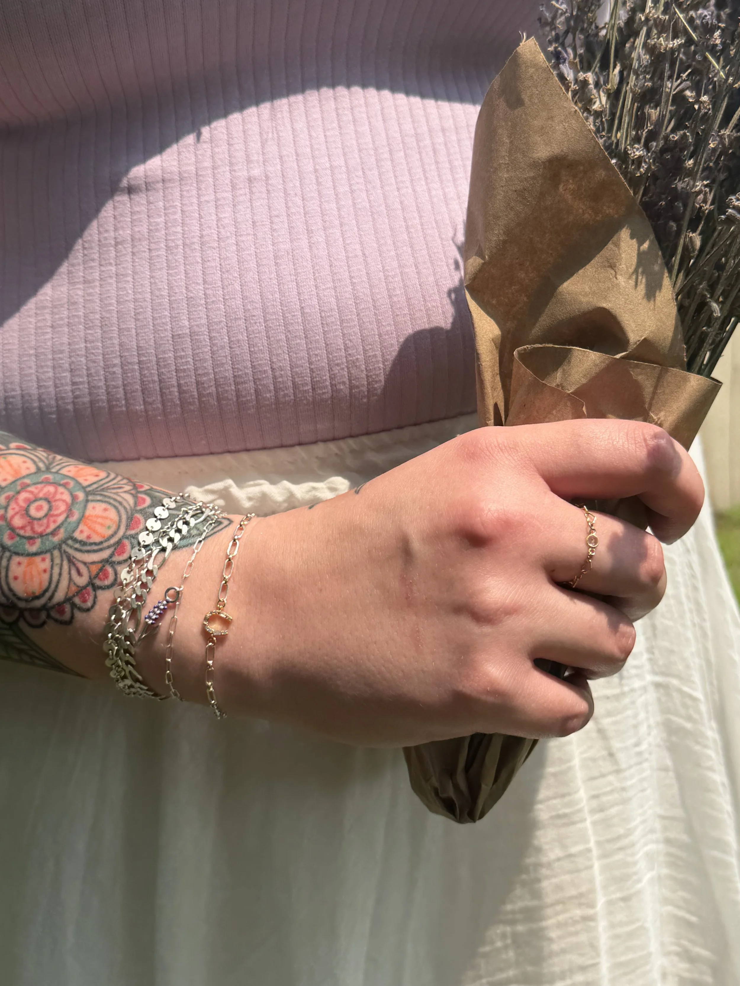 Close-up of a person holding a bouquet of dried lavender wrapped in brown paper, wearing a white textured skirt and multiple delicate silver bracelets on their wrist.