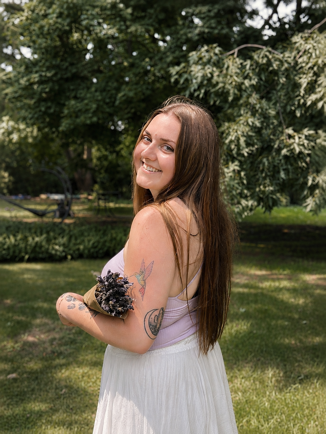 A young woman with long brown hair smiling outdoors in a park, with tattoos on her left arm, holding a bouquet of flowers.