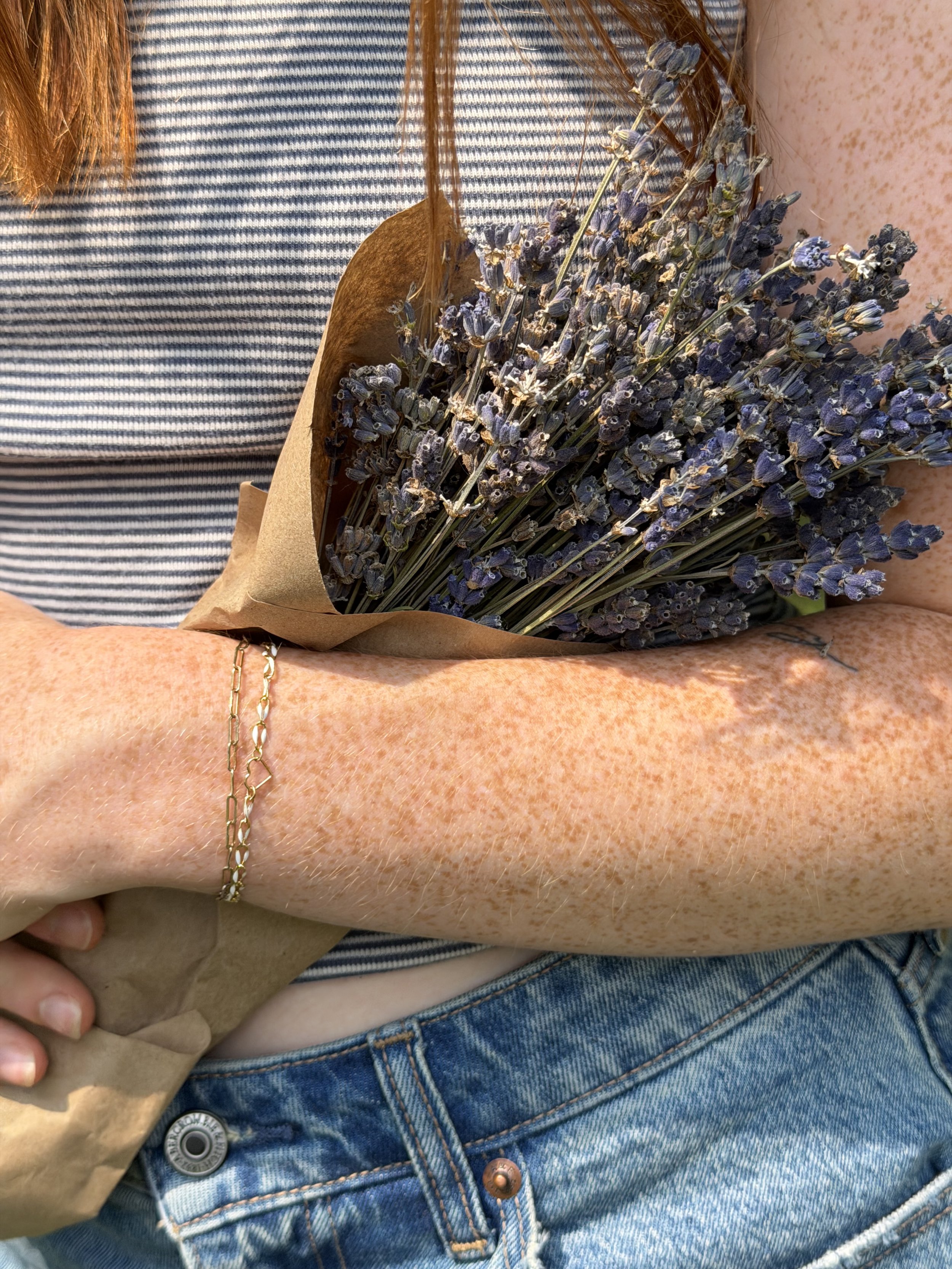 Person holding a bouquet of lavender flowers wrapped in brown paper, wearing a striped shirt, blue jeans, and a gold chain bracelet.