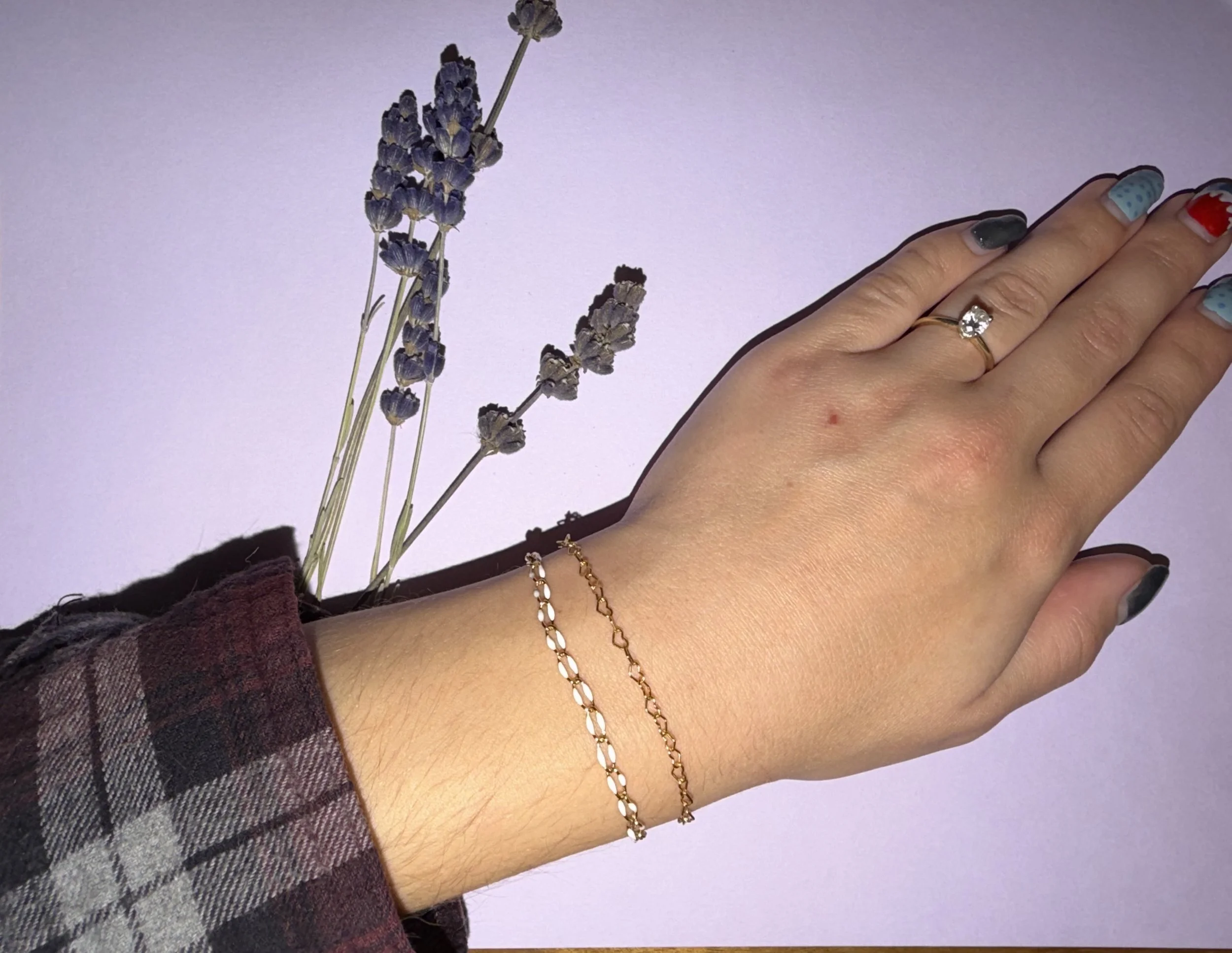 A person's hand and wrist with a gold ring and bracelets, wearing colorful nail polish, next to a sprig of lavender flowers on a light purple background.