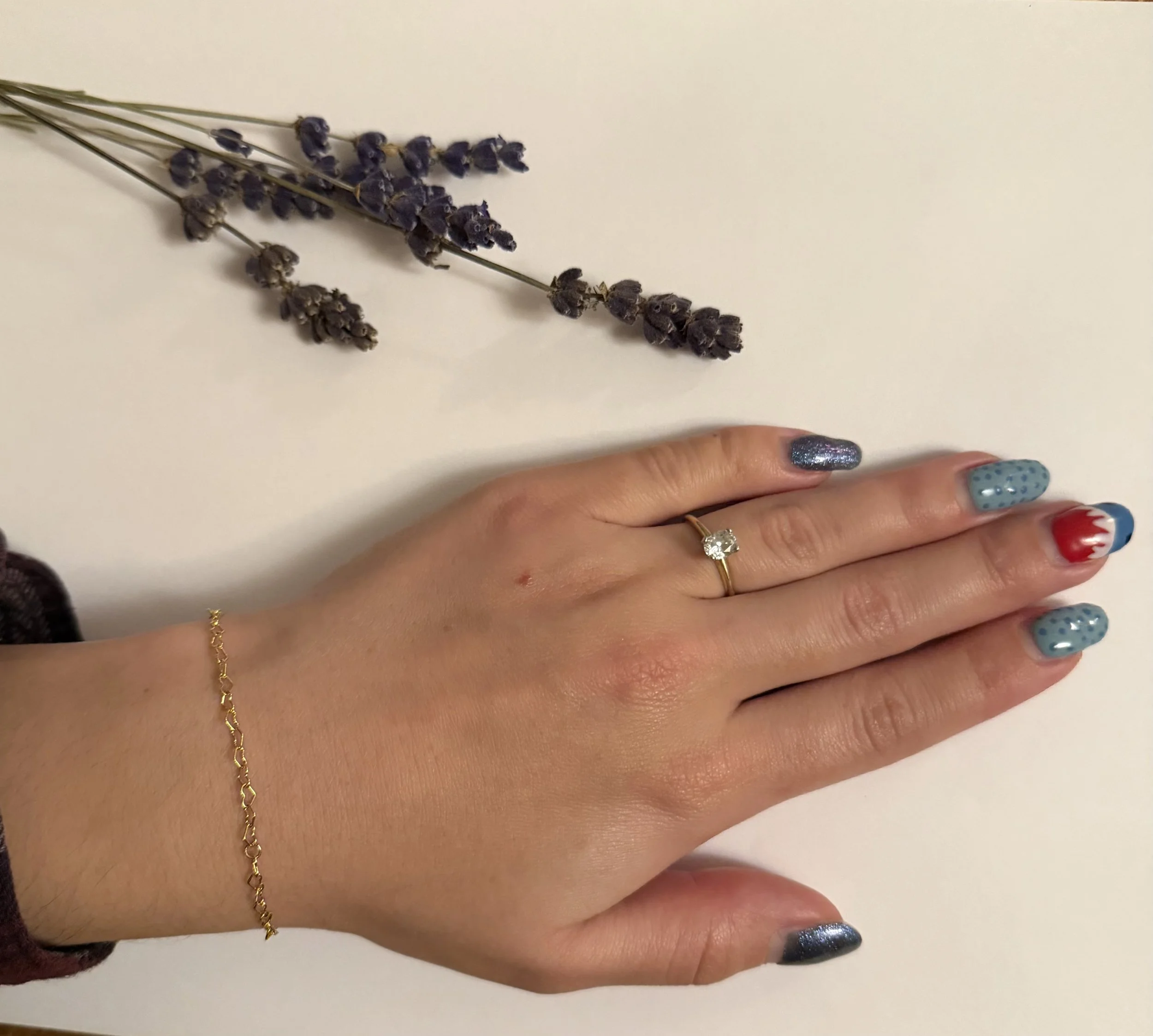 A hand with colorful American flag themed nail polish resting on a white surface, wearing a gold ring and a gold bracelet, with dried lavender flowers placed nearby.