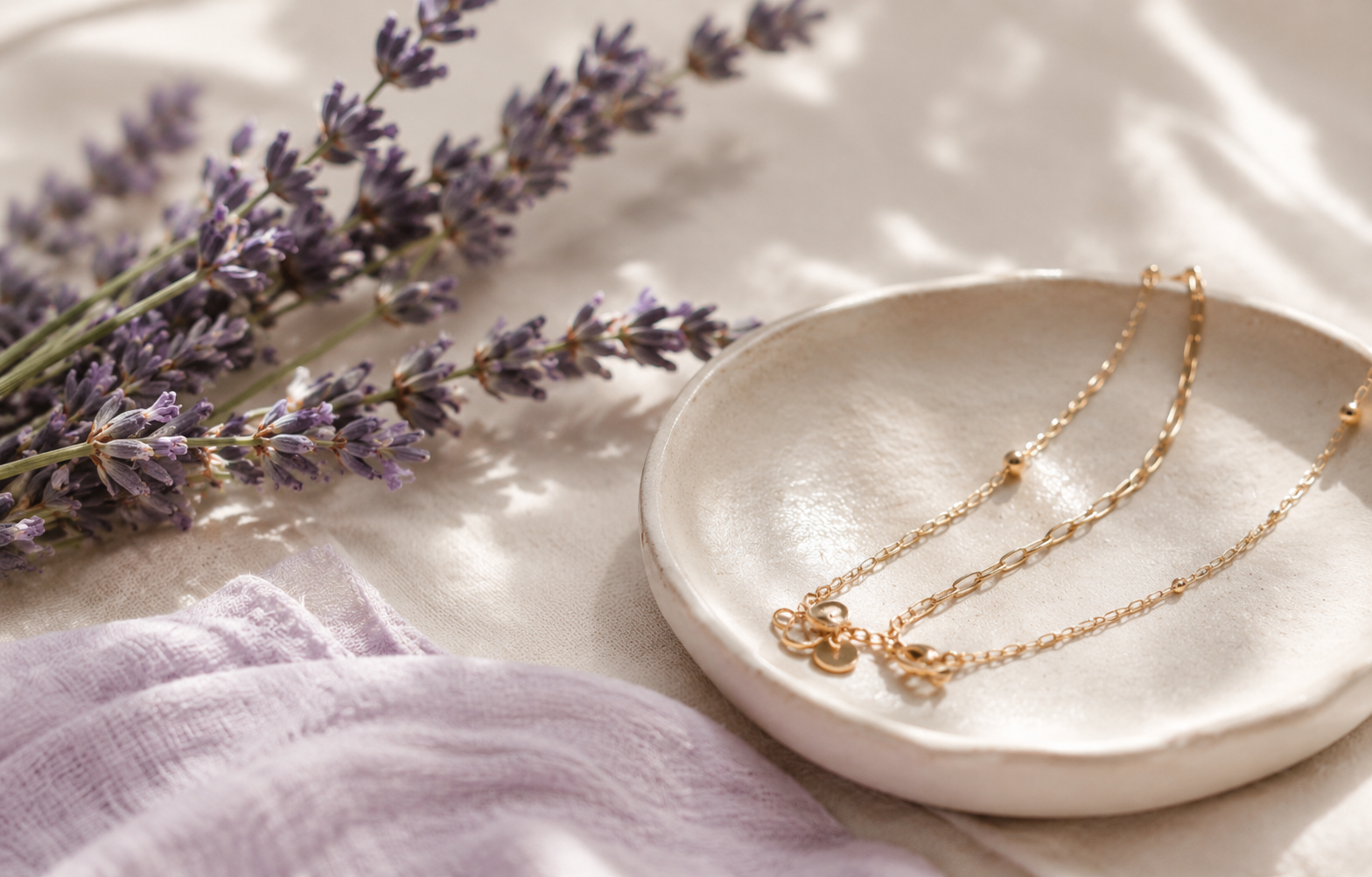 A white ceramic dish holding three gold necklaces with small pendants, with purple lavender flowers lying beside it on a soft fabric surface.