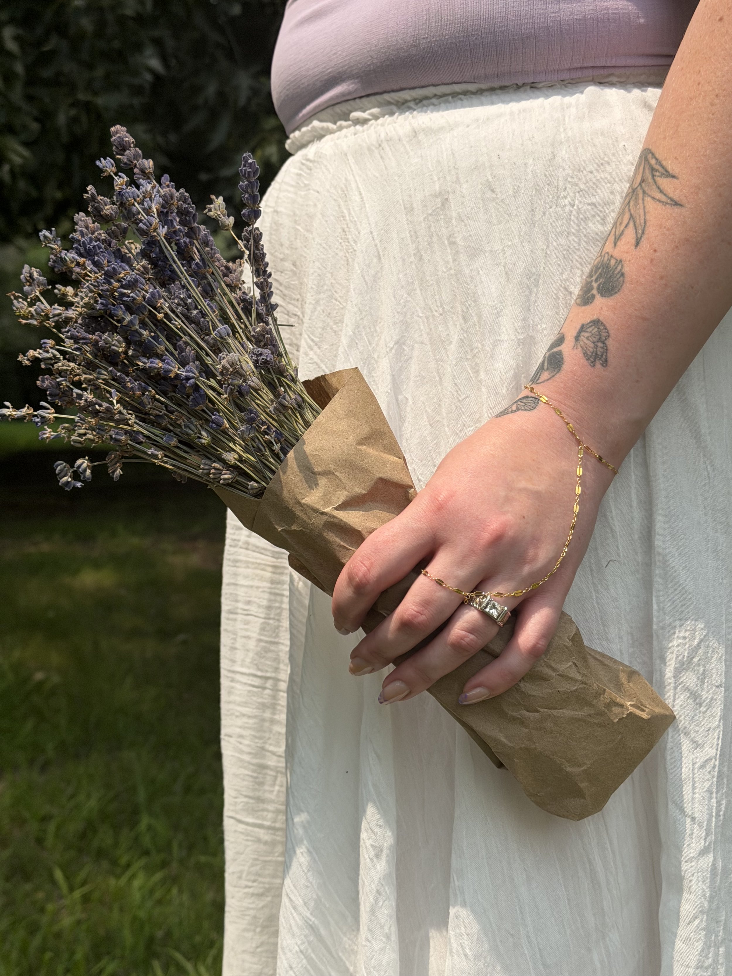 A person holding a bouquet of purple lavender flowers wrapped in brown paper, wearing a gold bracelet and a ring, with tattoos on their arm and wearing a light-colored skirt and top.