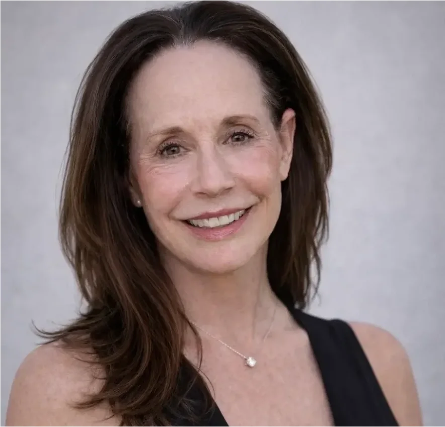 Portrait of a woman with long brown hair, smiling, wearing a black top and a necklace, against a light background.
