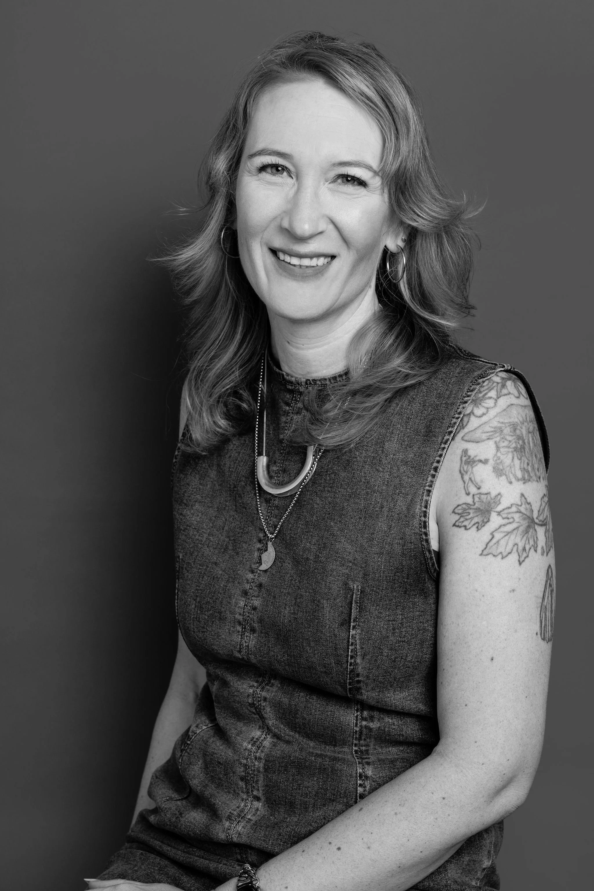 Black and white portrait of a woman with shoulder-length wavy hair, wearing hoop earrings, a necklace, and a sleeveless denim dress with tattoos on her right arm, smiling against a plain background.