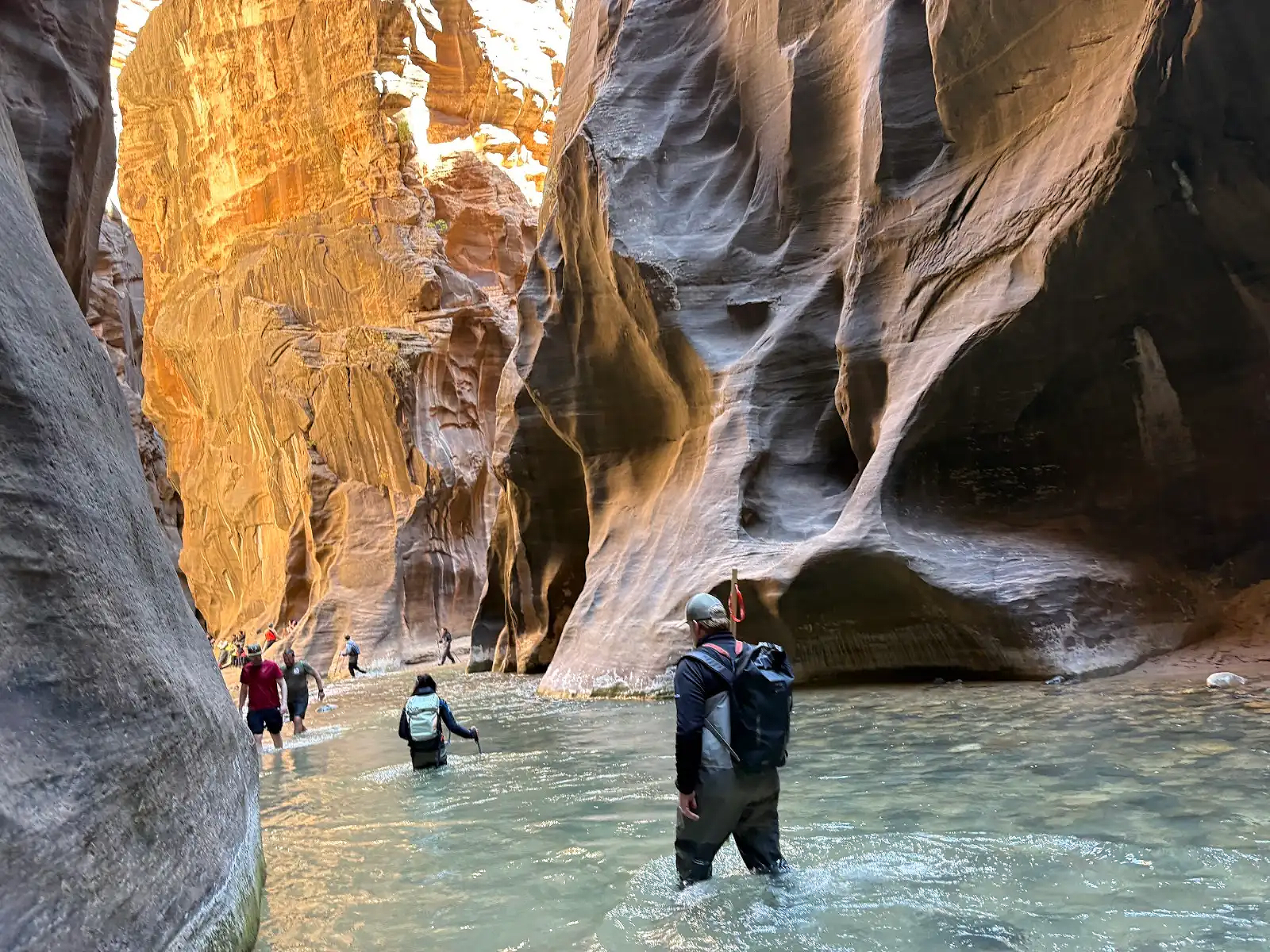 Hikers wading across a water-filled slot canyon with towering, smooth, colorful rock walls.