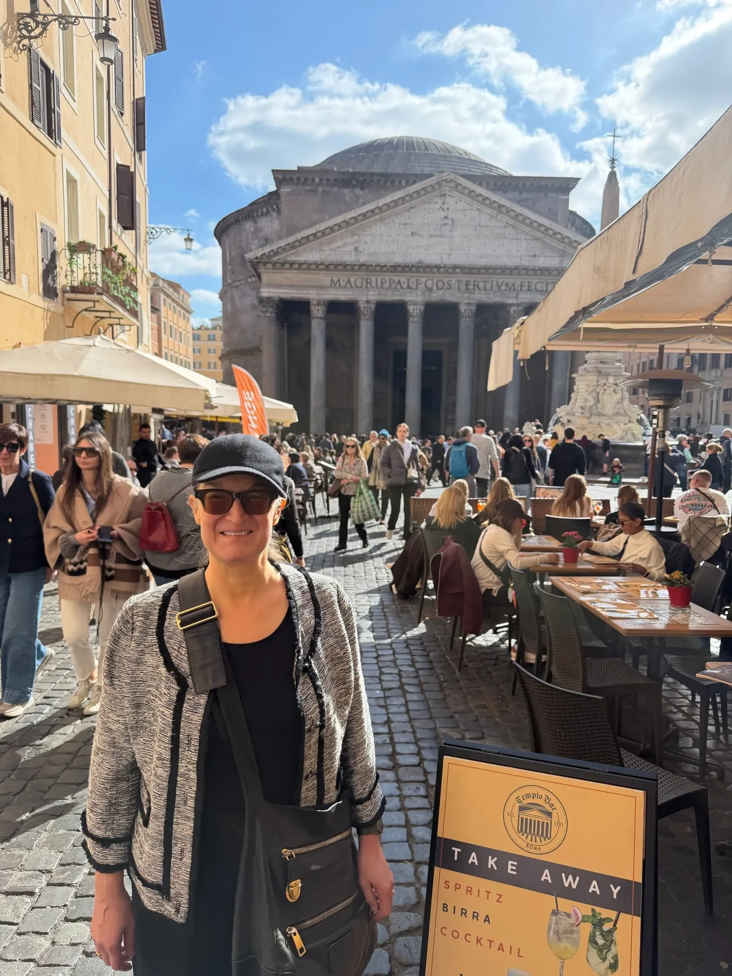 A woman wearing a hat and glasses in a cardigan, smiling in front of the busy town square in front of the Pantheon in Rome.