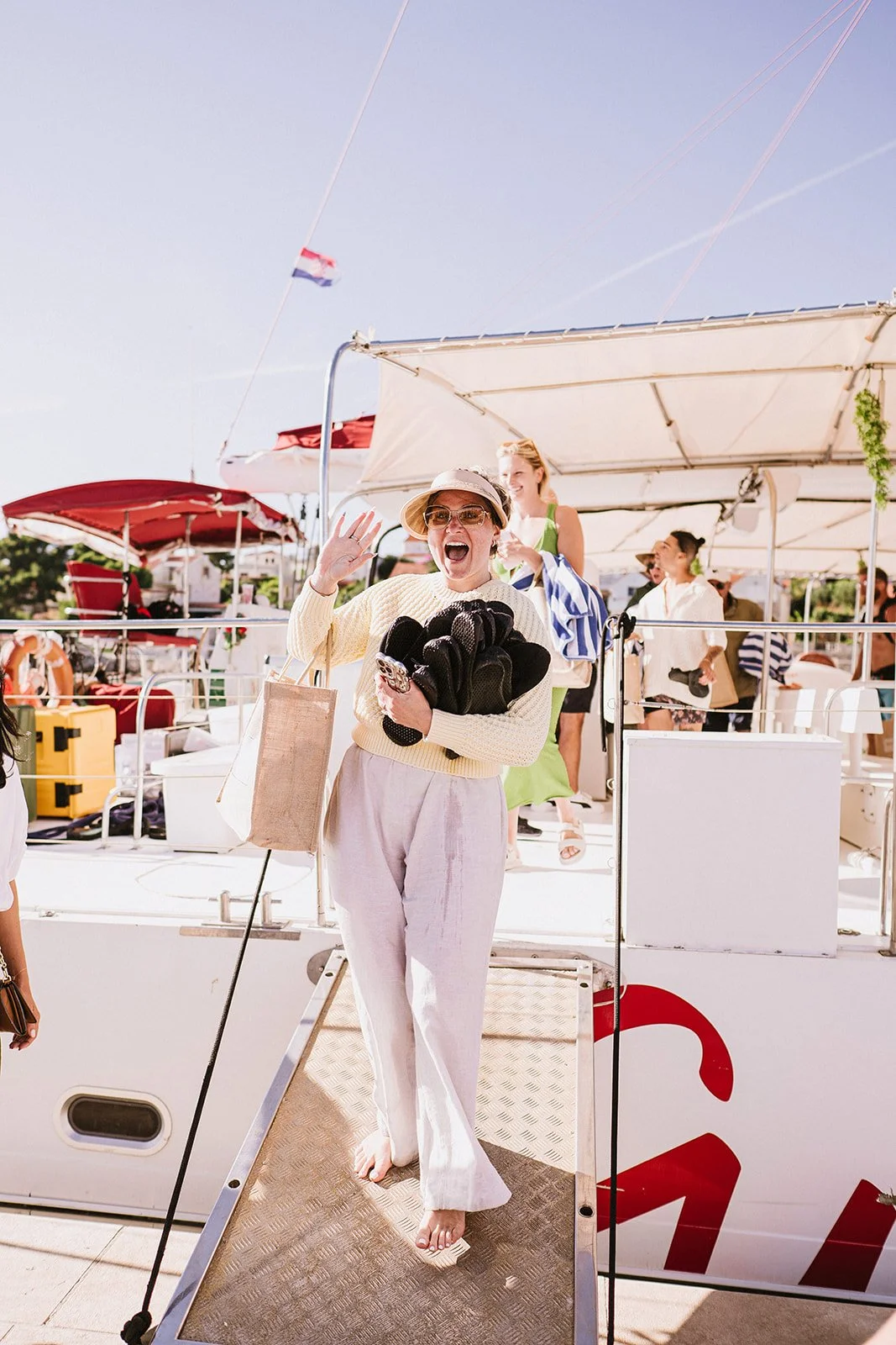 A woman in light-colored pants, a yellow sweater, and a sun hat waving and smiling on a boat, surrounded by other people and boats on a sunny day.