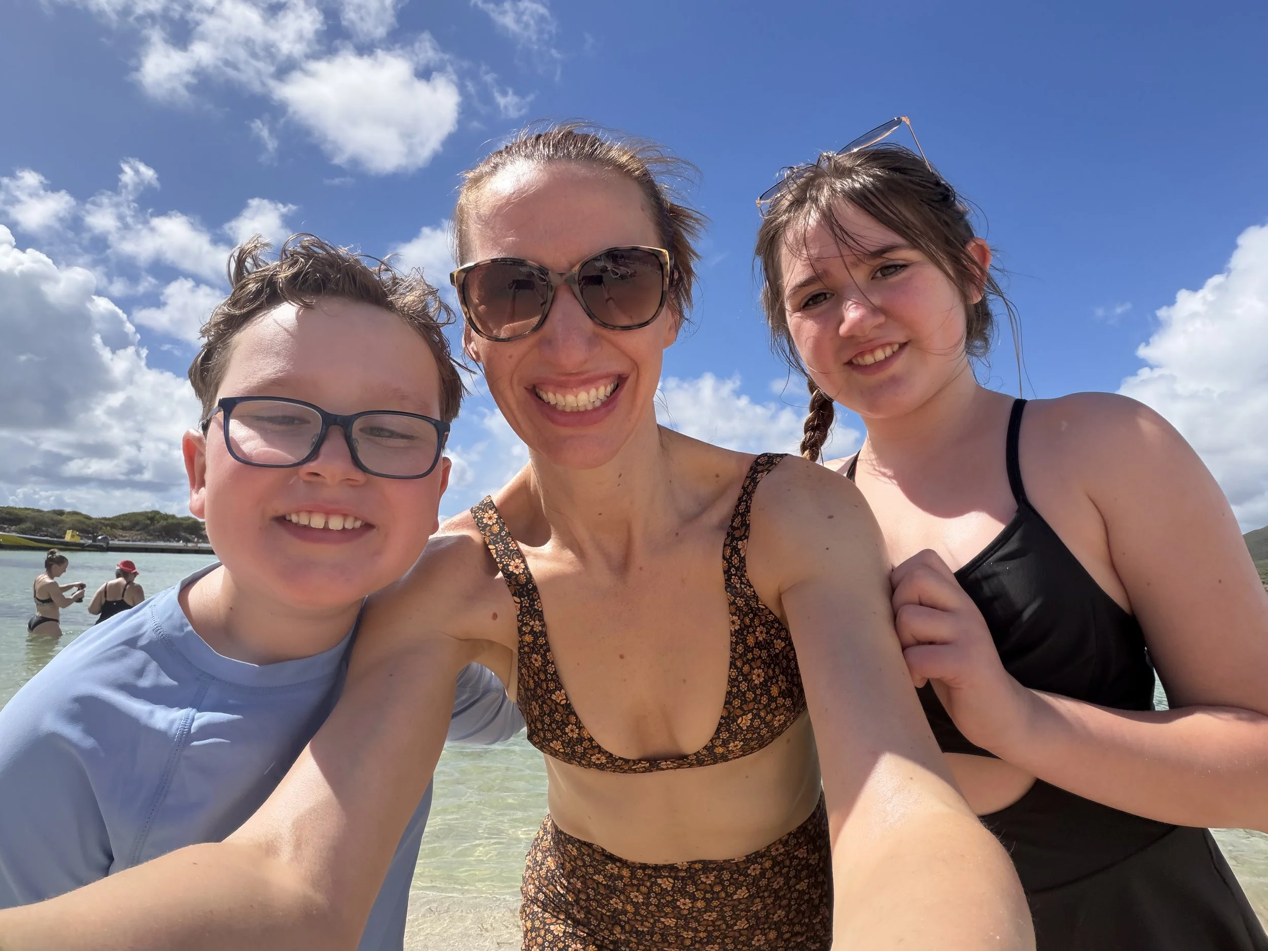 A mother taking a selfie with her son and daughter, everyone is in bathing suites and smiling with ocean and sky behind them.
