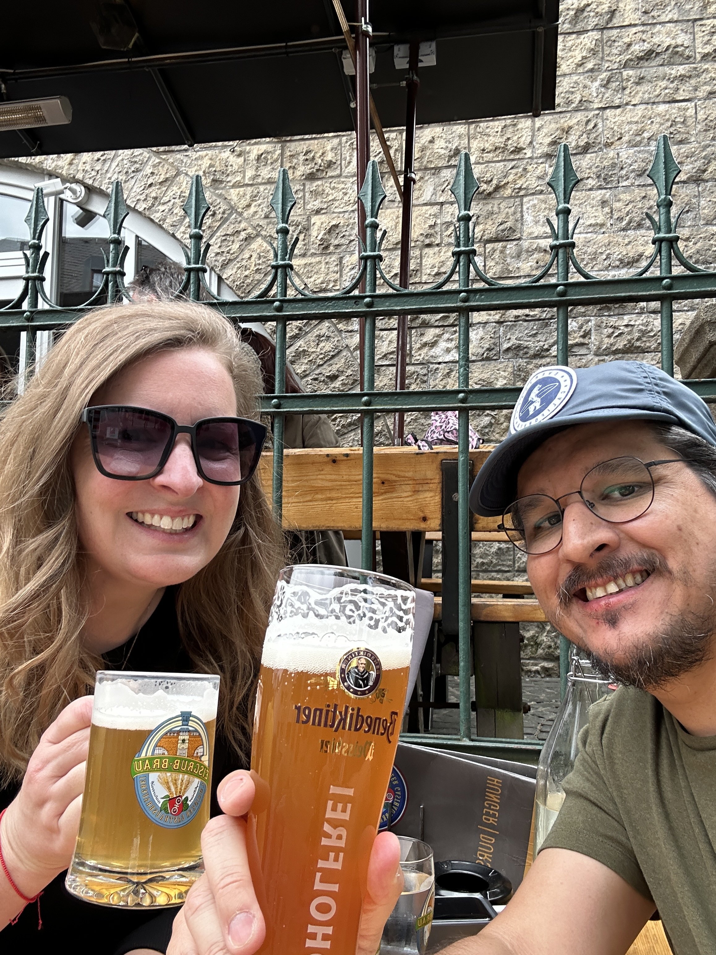 A woman with long hair and sunglasses and a man with a hat and glasses, cheering with big beer steins.
