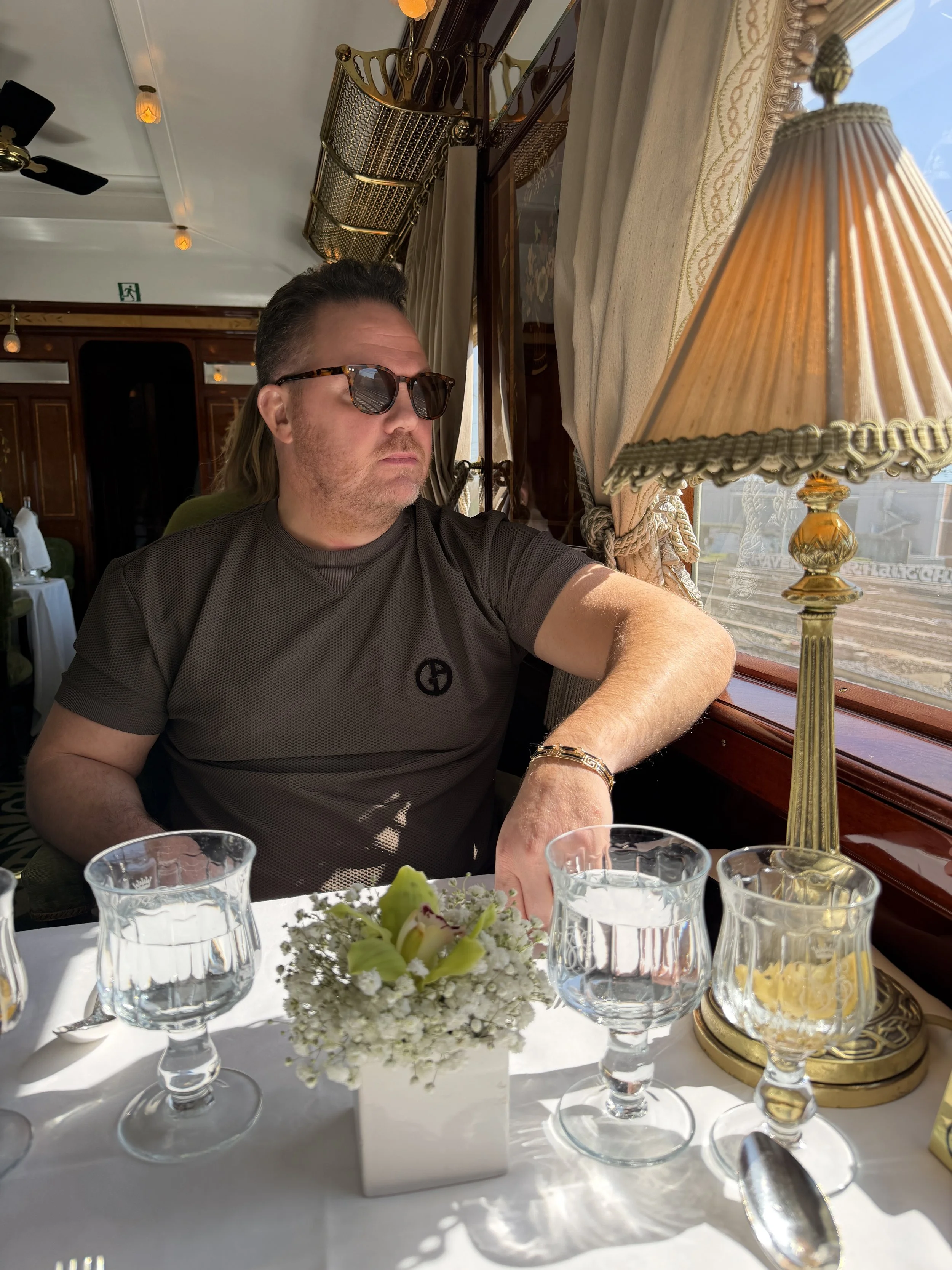 A man wearing sunglasses and looking out a train window with a vintage table lamp and several glasses of wine and water on the table in front of him.