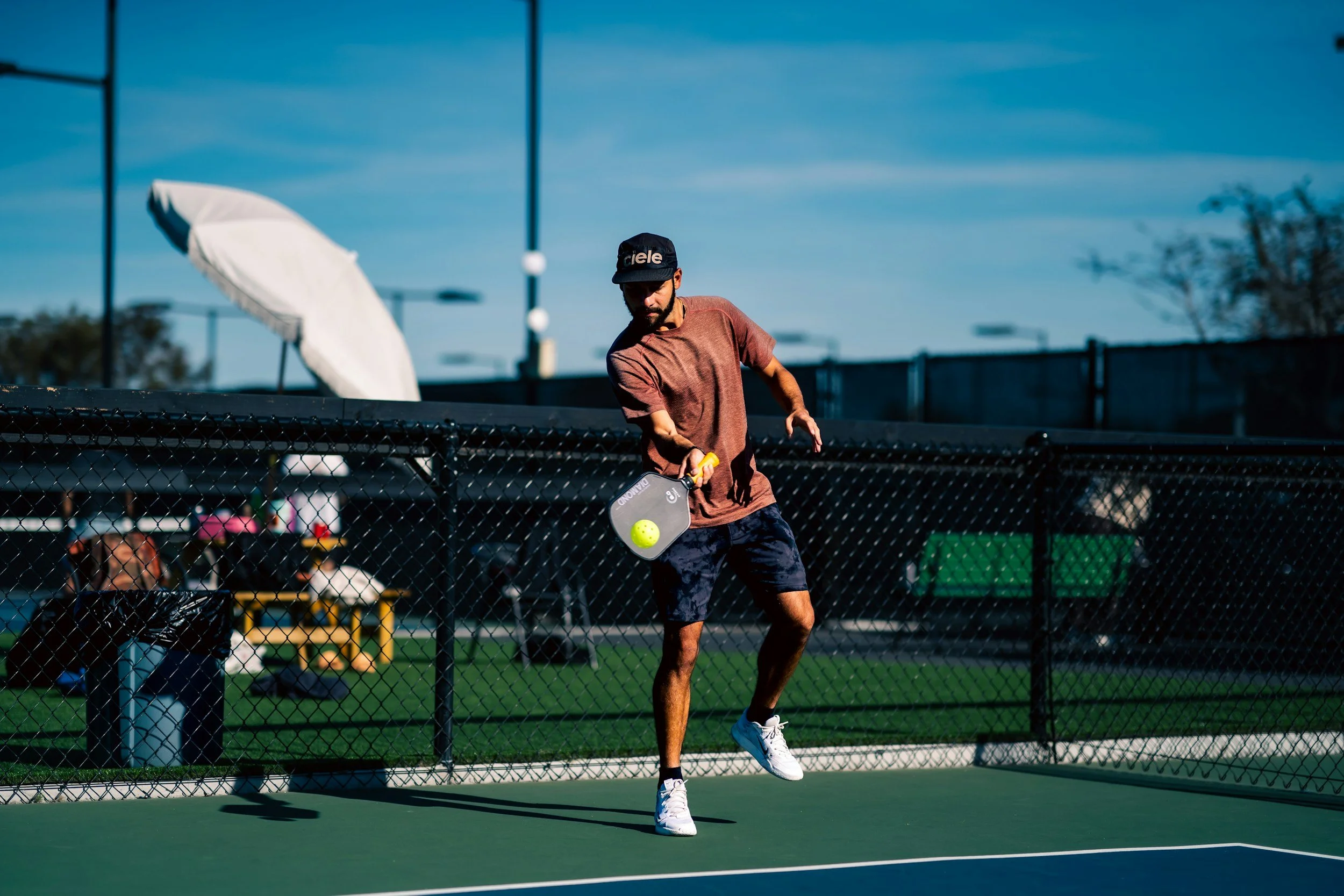 A man playing pickleball on an outdoor court, about to hit a yellow pickleball with a paddle, wearing a dark cap, T-shirt, and shorts, with a fence and equipment in the background.