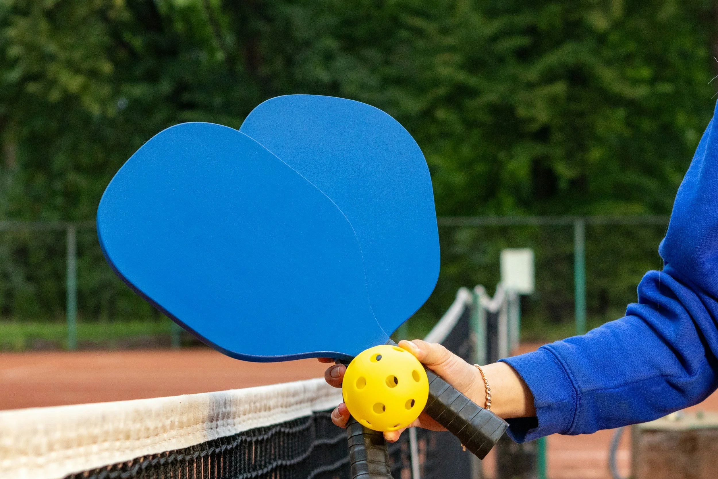 A person holding blue tennis paddles, a yellow ball, and wearing a blue jacket on an outdoor tennis court.