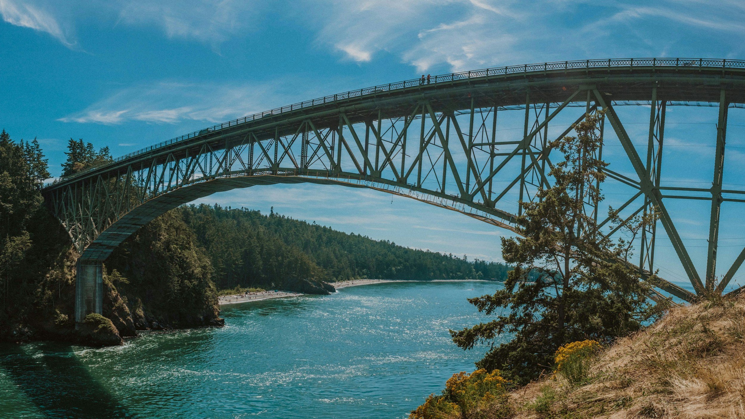 A large metal bridge arching over a body of water with trees on both sides, under a blue sky with some clouds.
