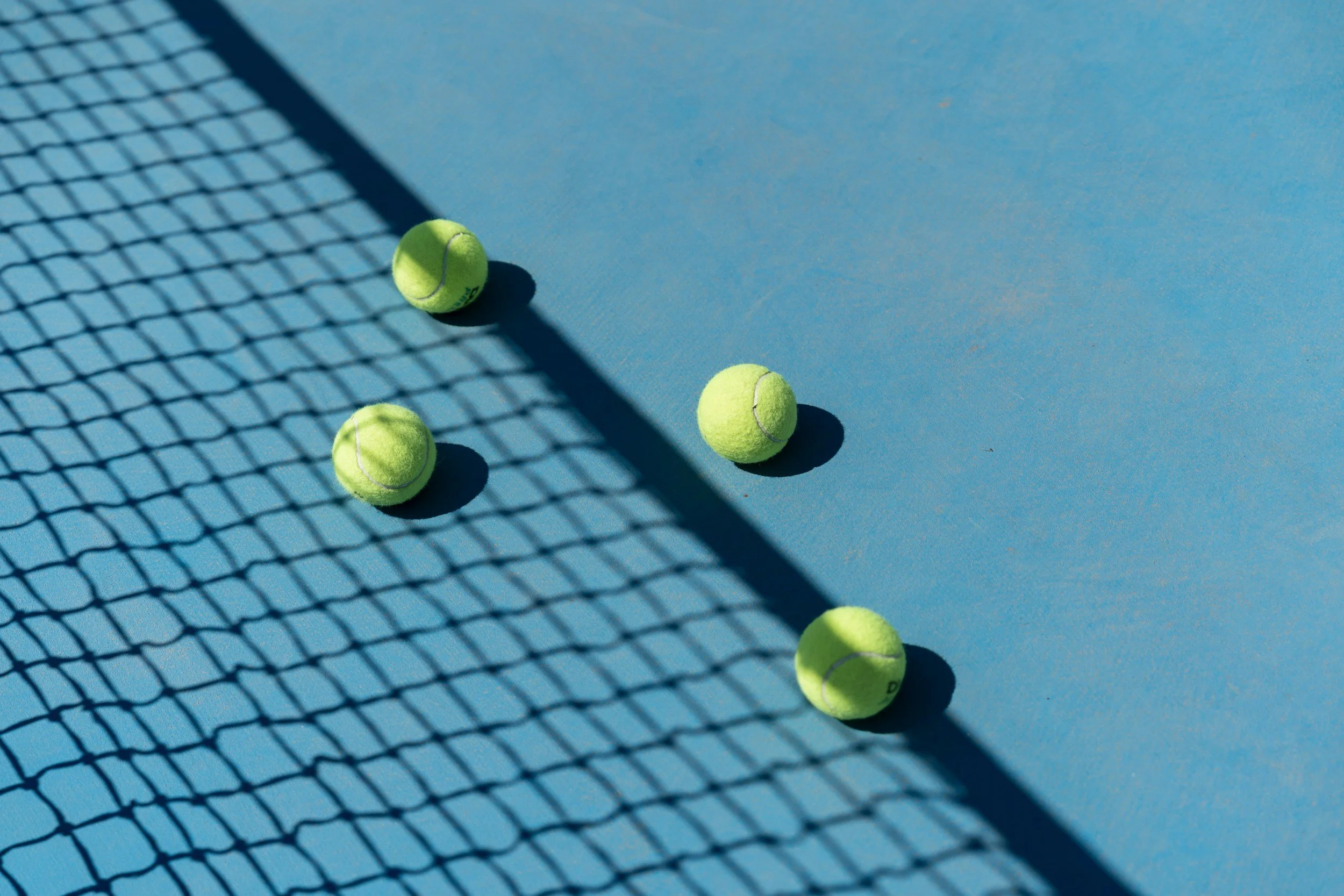 Several bright yellow tennis balls on a blue tennis court near a net, casting shadows.