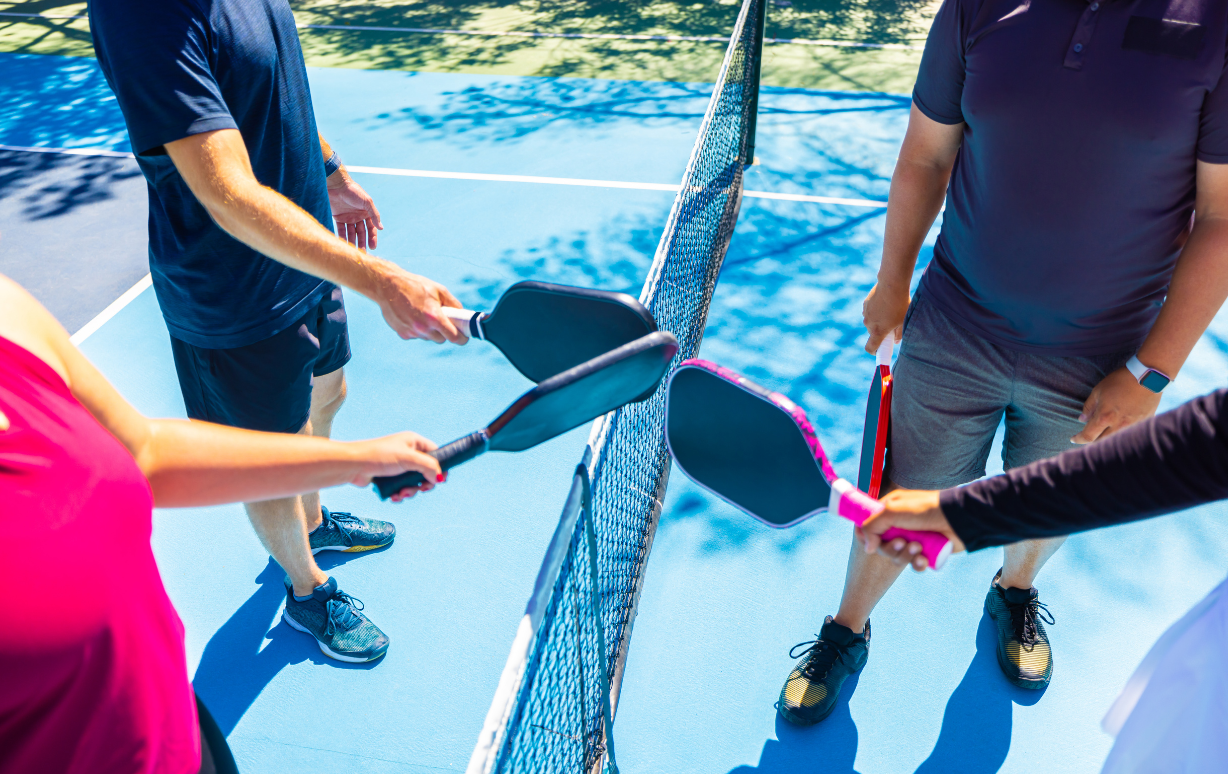People on an outdoor pickleball court holding paddles, about to hit a ball over the net.