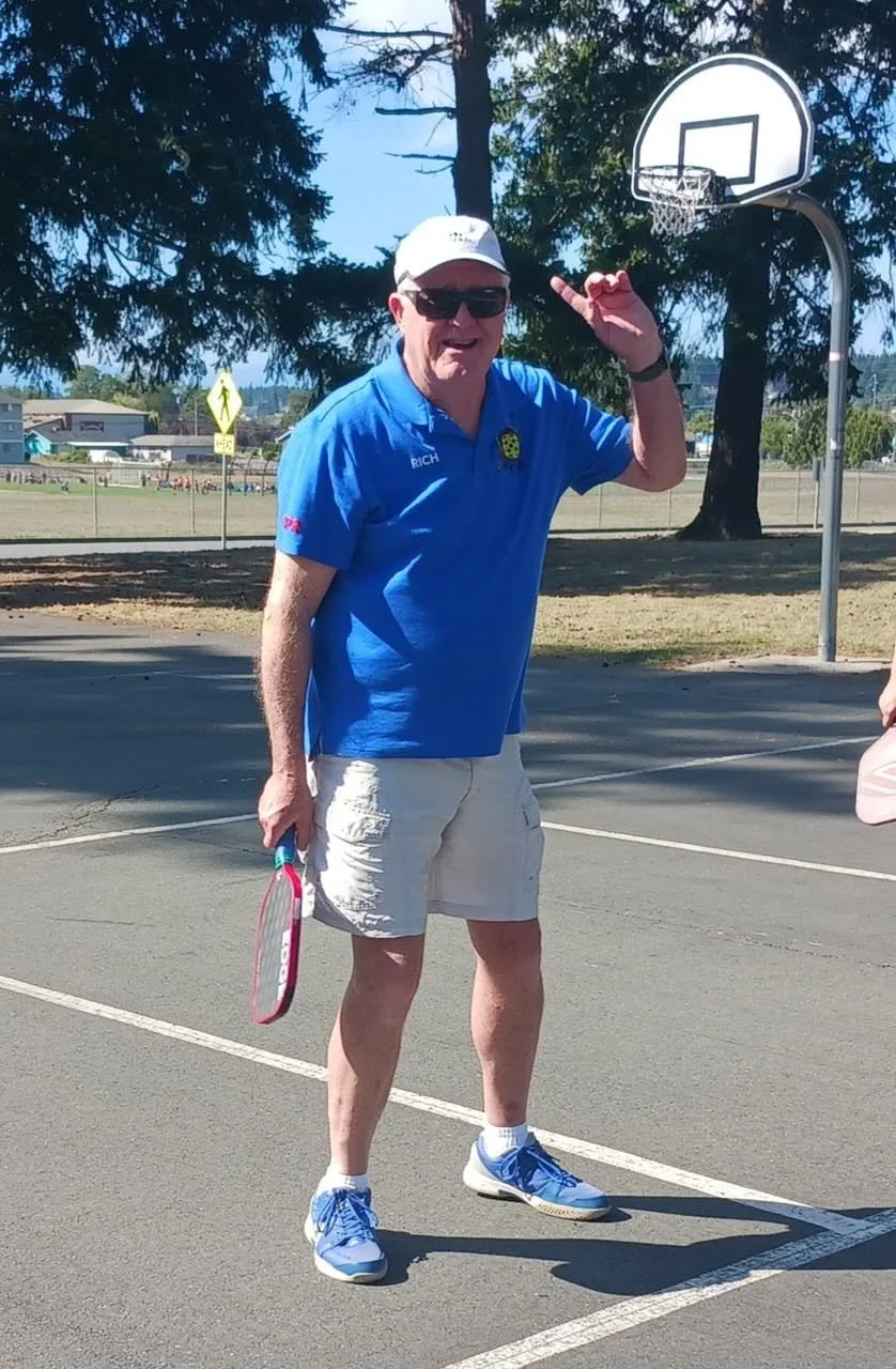 A man wearing sunglasses, a white cap, a blue polo shirt, khaki shorts, and blue sneakers standing on a basketball court with a basketball in his left hand and making a gesture with his right hand near a basketball hoop.